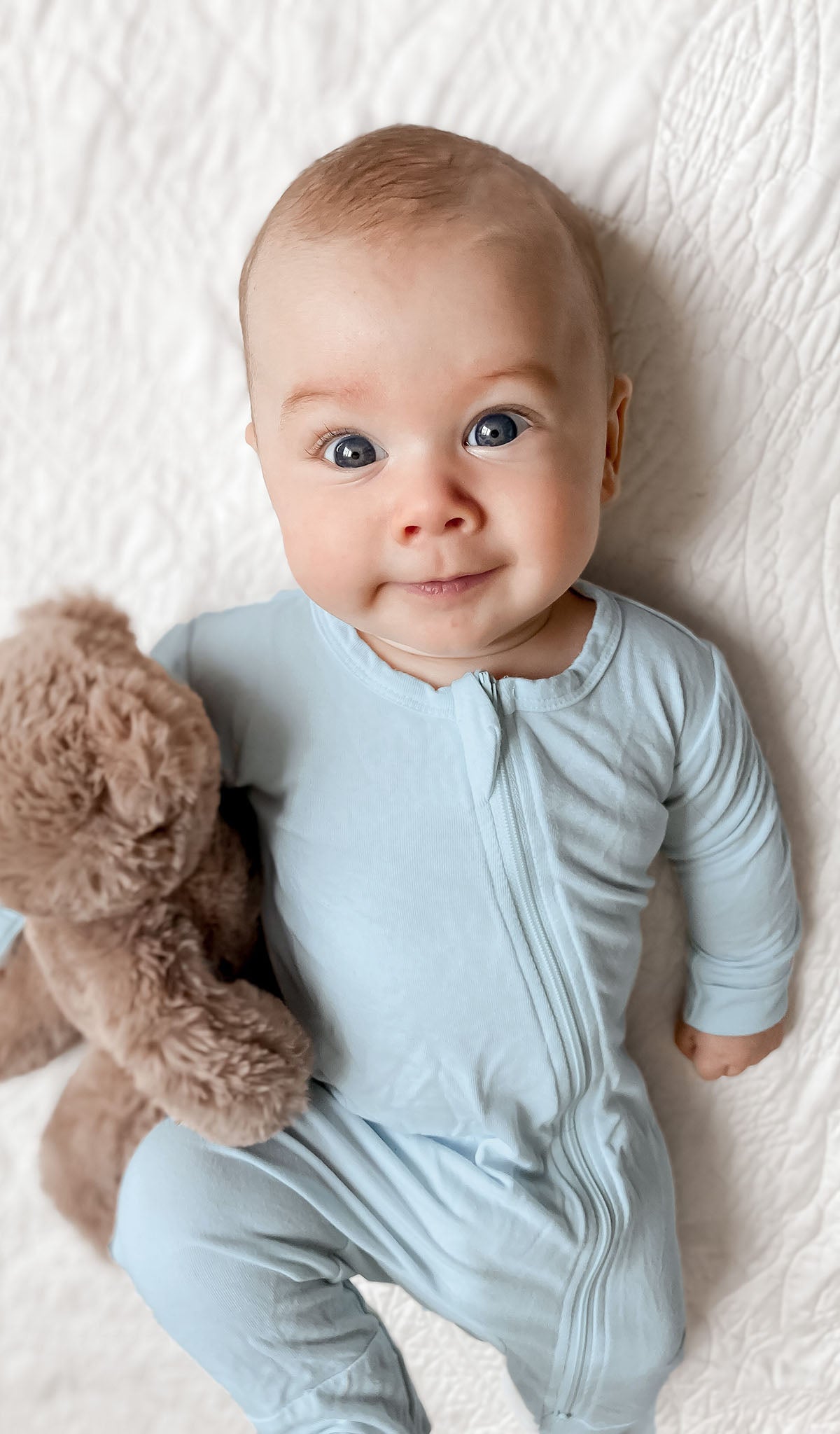 Whispering Blue Footie worn by baby holding teddy bear while laying on a quilt.