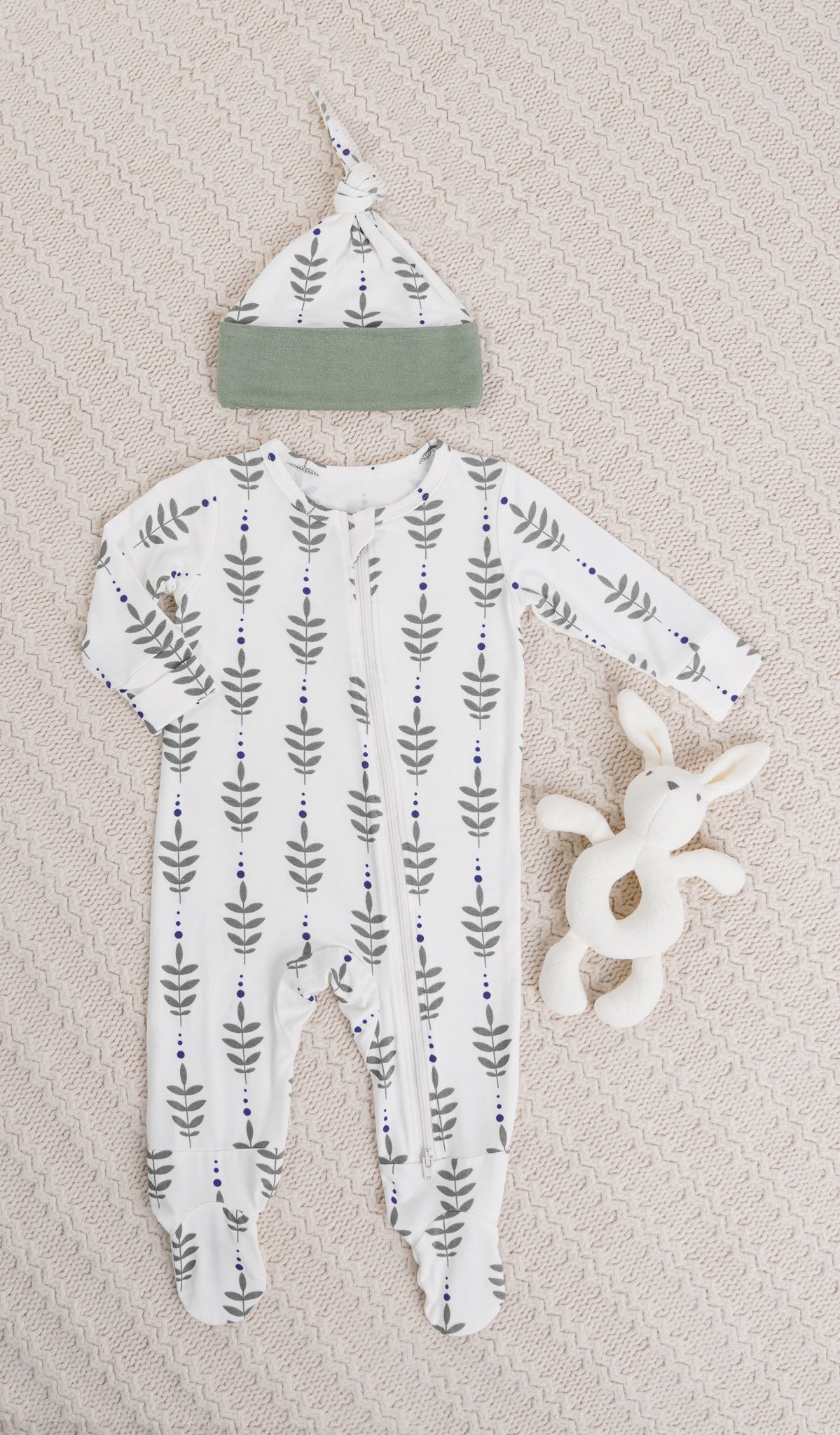 Eucalyptus Footie 2-Piece. Long sleeve footie with zip front, matching knotted baby hat and bunny rattle on knitted beige blanket.