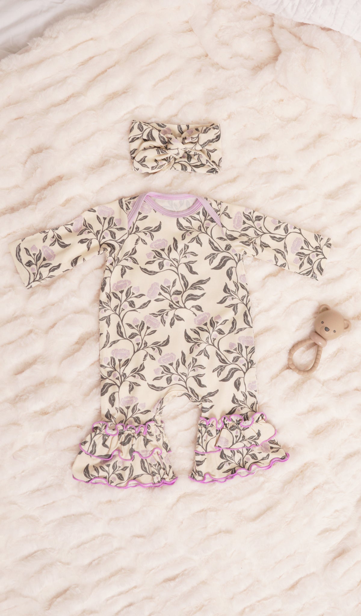 Poppy Ruffle Romper 2-Piece. Long sleeve romper with ruffles on legs with matching headwrap and a rattle on soft beige bedding.