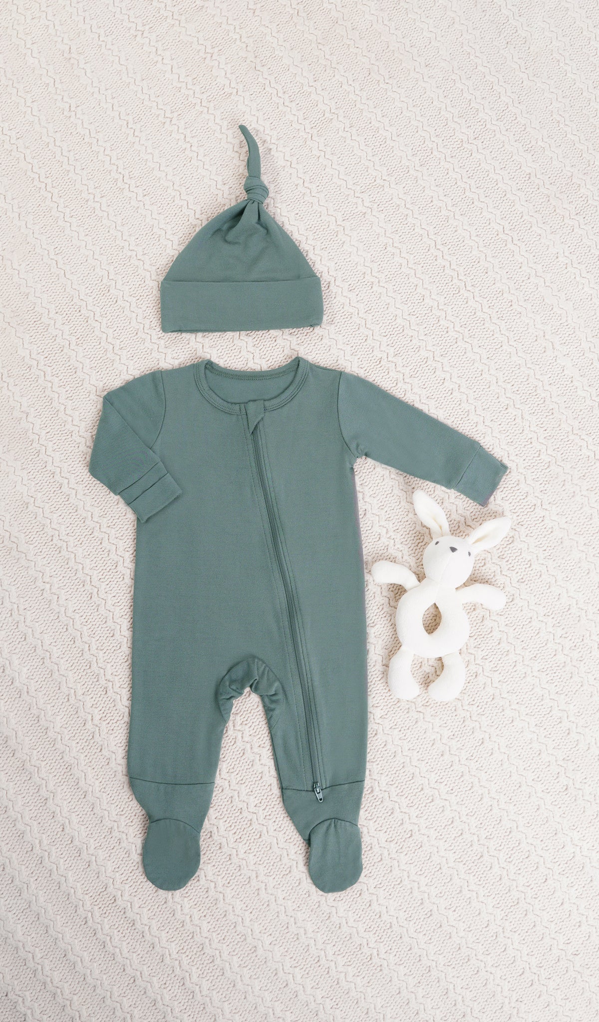 Fog Footie 2-Piece. Long sleeve footie with zip front, matching knotted baby hat and a bunny rattle on knitted beige blanket.