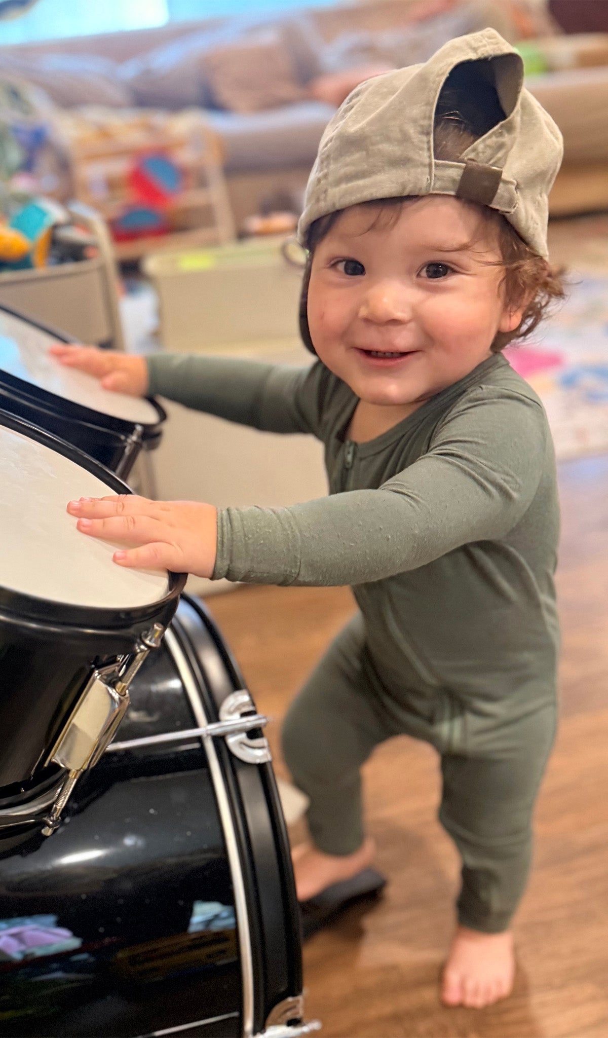 Baby wearing convertible romper with hat turned backwards and playing with drum set while standing.