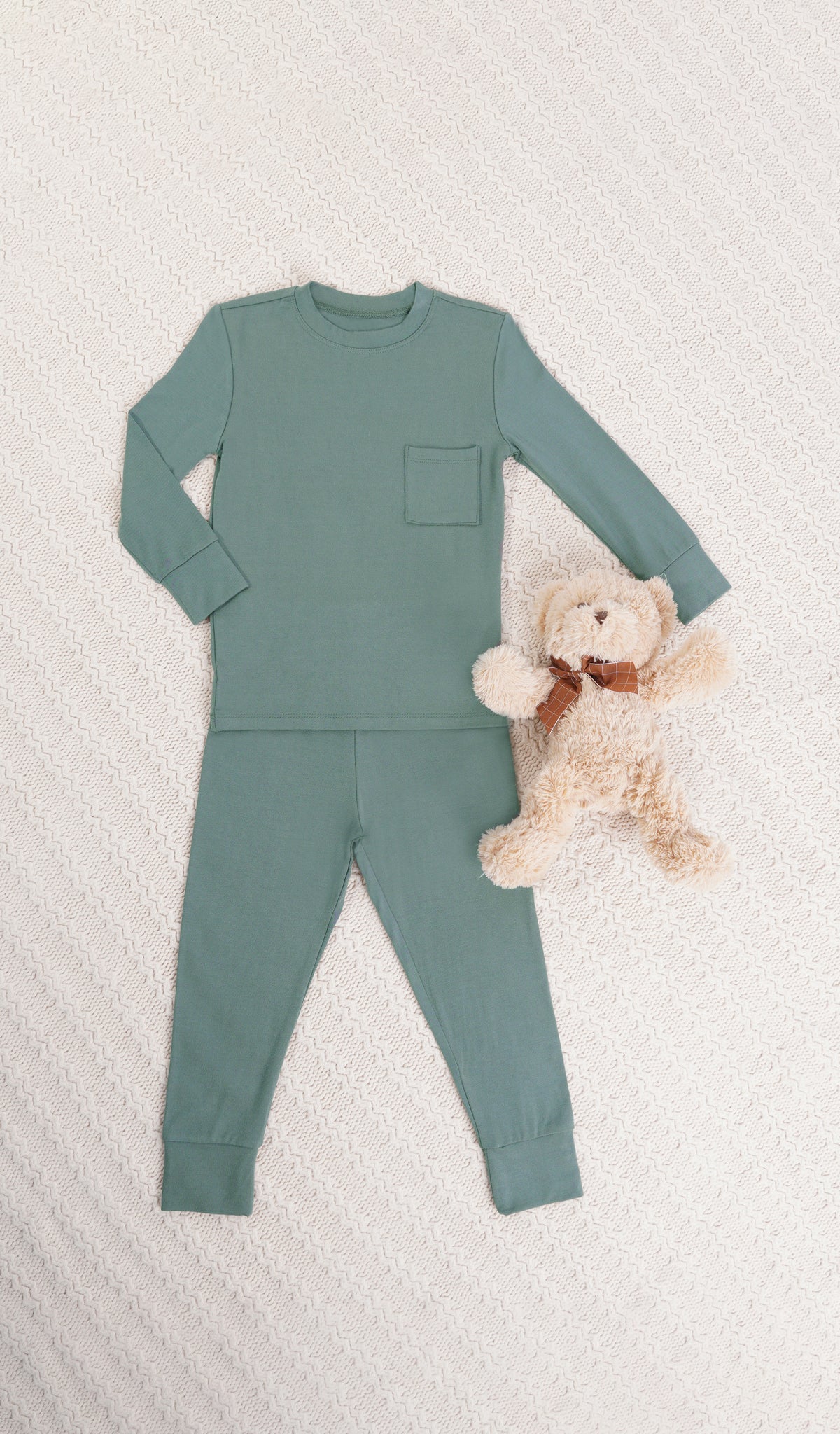 Fog Emerson Kids 2-Piece Pant PJ. Long sleeve top with cuff trim and long pant with cuff trim and a stuffed bear on a knitted beige blanket.
