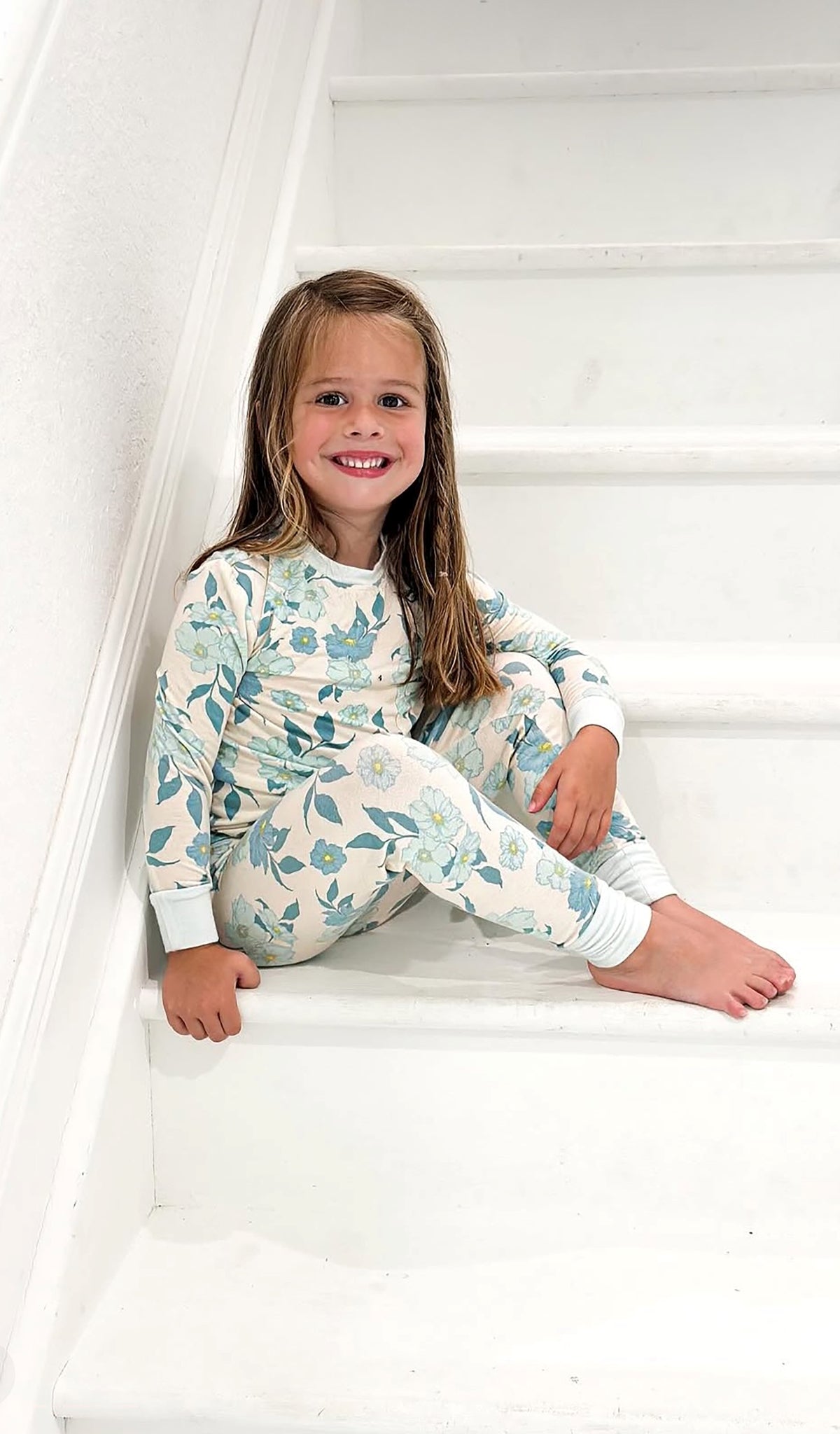A young girl with long light brown hair sits smiling on white wooden stairs, wearing a long-sleeve pajama set with a light blue floral and leaf pattern on a cream background.