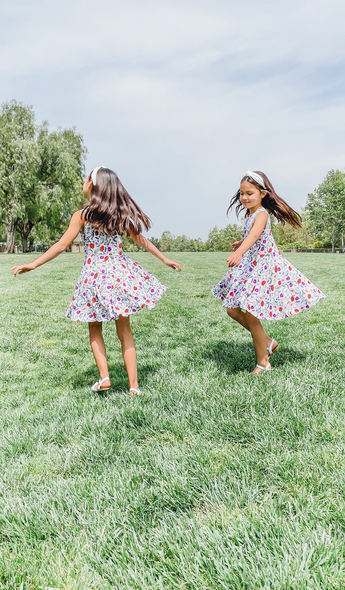 Lucia Kids Twirly Dress in Zinnia worn by two sisters twirling on a grass field.