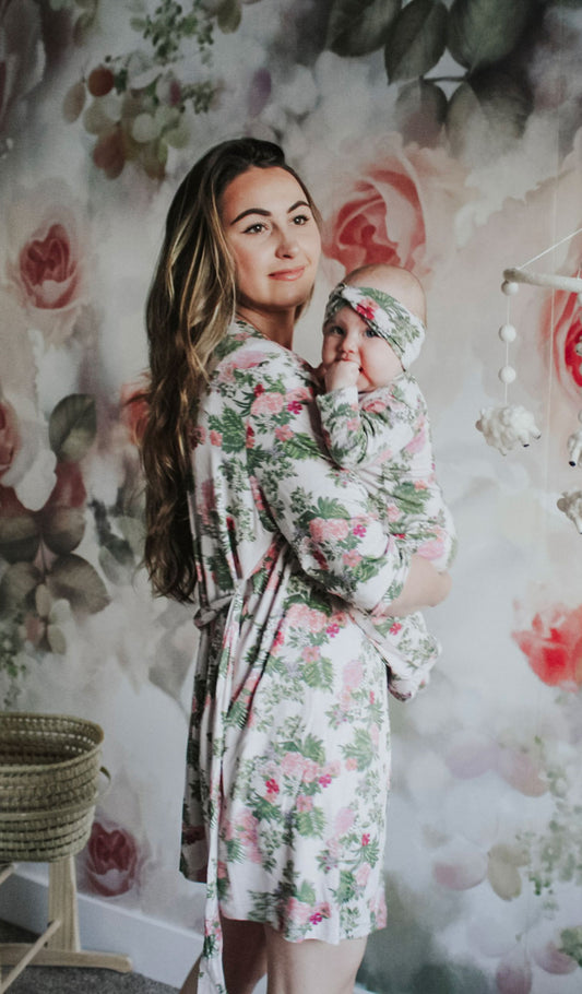 Beige Floral Adalia 5-Piece Set. Lifestyle shot of woman standing wearing robe and short while holding baby girl wearing matching baby footie and headband.