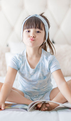 Blue Chantilly Bella Kids 3-Piece Short PJ. Little girl making a kissing face, sitting on bed with book wearing short sleeve top and short with matching headwrap.