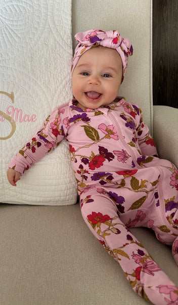 Lavender Rose Footie 2-Piece Set. Smiling baby wearing zip front footie with matching headwrap tied into a tie-knot bow, while sitting on a chair.
