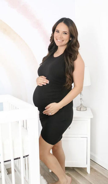 Black Tamara dress worn by pregnant woman with two hands on her belly and standing next to a crib.
